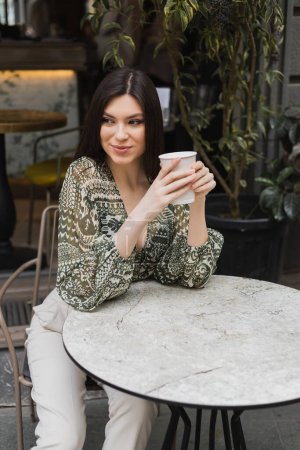 young woman with long brunette hair sitting on chair near round bistro table and holding paper cup with drink while looking away and smiling near blurred plants on terrace of cafe in Istanbul