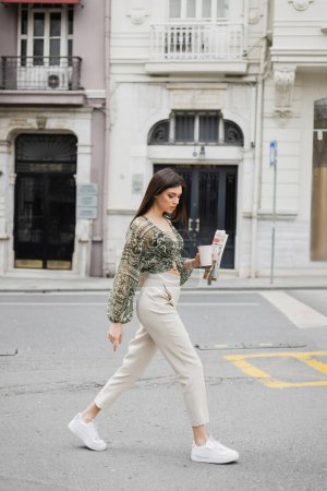hübsche junge Frau mit langen brünetten Haaren und Make-up, die Pappbecher mit Kaffee und Zeitung hält, während sie in trendigem Outfit mit beiger Hose und Bluse auf einer städtischen Straße in der Nähe eines Gebäudes in Istanbul spaziert 