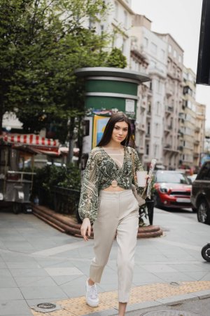 pretty woman with long hair and makeup holding paper cup with coffee and newspaper while walking in trendy outfit with handbag on chain strap on street near blurred building and cars in Istanbul 