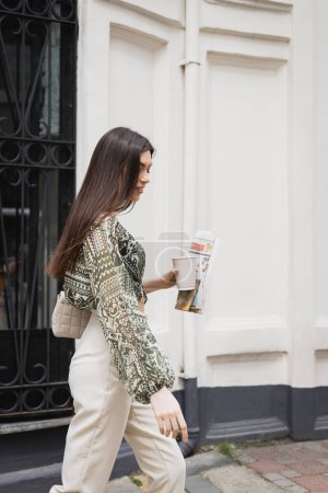 mujer sonriente con pelo largo morena y maquillaje sosteniendo taza de papel con café y periódico mientras camina en traje de moda y bolso en la calle urbana cerca de valla metálica y pared blanca en Estambul 