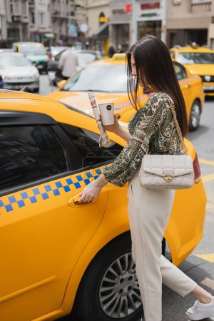 schicke Frau mit langen Haaren, die Pappbecher mit Kaffee und Zeitung in der Hand hält, während sie im trendigen Outfit mit Handtasche am Kettenriemen in der Nähe des gelben Taxis auf einer verschwommenen Stadtstraße in Istanbul steht 