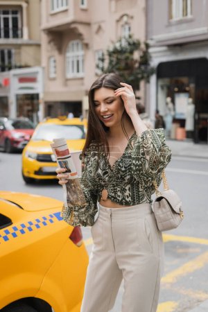 szczęśliwa kobieta dostosowując swoje długie włosy i trzymając papierowy kubek z kawą i gazetą stojąc w modnym stroju z torebką na pasku łańcucha w pobliżu żółtej taksówki na rozmytej ulicy miejskiej w Stambule 