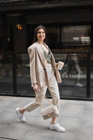 lächelnde Frau mit langen Haaren, die im Blazer mit beiger Hose, abgeschnittener Bluse und Handtasche am Kettenriemen spaziert und in der Nähe eines Cafés in der Stadtstraße in Istanbul Pappbecher mit Kaffee hält 
