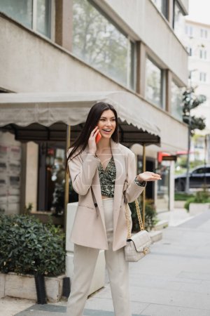 Photo for Happy young woman with brunette long hair and makeup smiling while talking on smartphone and standing in trendy outfit with handbag on chain strap near blurred fancy restaurant in Istanbul - Royalty Free Image