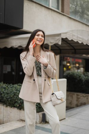 hermosa mujer con cabello largo morena y maquillaje sonriendo mientras habla en el teléfono inteligente y de pie en traje de moda con bolso de mano en correa de cadena cerca de un restaurante de lujo borroso en Estambul 
