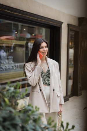 stylische junge Frau mit brünetten langen Haaren und Make-up im Smartphone-Gespräch und im trendigen Outfit mit Handtasche am Kettenriemen in der Nähe von verschwommenem Juweliergeschäft und Werk in Istanbul 