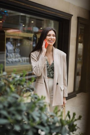 smiling young woman with brunette long hair and makeup talking on smartphone and standing in trendy outfit with handbag on chain strap near blurred jewelry store and plant in Istanbul 