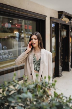 surprised young woman with brunette long hair and makeup talking on smartphone and standing in trendy outfit with handbag on chain strap near blurred jewelry store and plant in Istanbul 