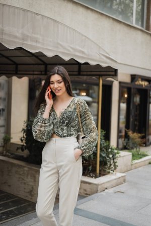 stylish young woman with brunette long hair and makeup smiling while talking on smartphone and standing in trendy outfit with handbag on chain strap near blurred fancy restaurant in Istanbul 