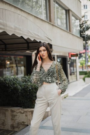 stylish young woman with brunette long hair and makeup smiling while talking on smartphone and standing in trendy outfit with handbag on chain strap near blurred fancy restaurant in Istanbul 