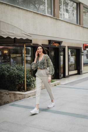 amazed young woman with brunette long hair and makeup smiling while talking on smartphone and walking in trendy outfit with handbag on chain strap near blurred fancy restaurant in Istanbul 