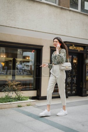 cheerful young woman with long hair and makeup smiling while talking on smartphone and walking in trendy outfit with handbag on chain strap near blurred jewelry store in Istanbul 