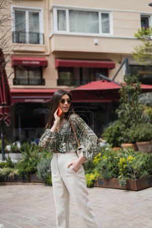 junge Frau mit langen Haaren und Sonnenbrille, die lächelt, während sie mit dem Smartphone spricht und mit der Hand in der Tasche und der Handtasche am Kettenriemen in der Nähe verschwommener Gebäude und Anlagen in Istanbul steht 