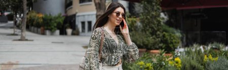 cheerful young woman with long hair and sunglasses smiling while talking on smartphone and walking with handbag on chain strap near blurred building and plants on street in Istanbul, banner 