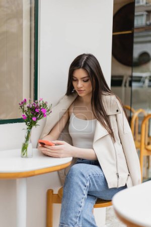 mujer joven con el pelo largo sentado en la silla cerca de la mesa del bistro con flores en jarrón y mensajes de texto en el teléfono inteligente mientras está sentado en ropa de moda con chaqueta de cuero en la cafetería en la terraza al aire libre en Estambul 