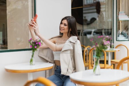 mujer feliz con el pelo largo sentado en la silla cerca de la mesa del bistro con flores en jarrón y tomar selfie en el teléfono inteligente mientras posa en ropa de moda en la cafetería en la terraza al aire libre en Estambul 