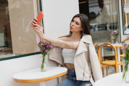 woman with long hair sitting on chair near bistro table with flowers in vase and pouting lips, taking selfie on smartphone while posing in trendy clothes in cafe on terrace outdoors in Istanbul 