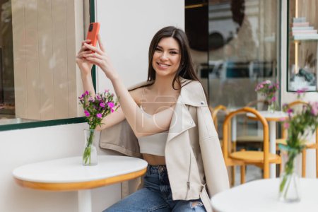 mujer sonriente con el pelo largo sentado en la silla cerca de la mesa del bistro con flores en jarrón y tomar selfie en el teléfono inteligente mientras posa en ropa de moda en la cafetería en la terraza al aire libre en Estambul 