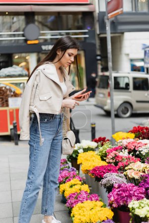 mujer morena con el pelo largo de pie en chaqueta de cuero beige, vaqueros y bolso con correa de cadena que sostiene el teléfono inteligente mientras mira las flores cerca de coches borrosos en la calle en Estambul, vendedor