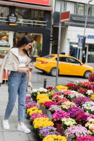 mujer morena con pelo largo de pie en chaqueta de cuero beige, vaqueros y bolso con correa de cadena que sostiene el teléfono inteligente cerca de las flores junto a la calle borrosa en Estambul, vendedor