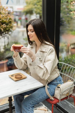 glückliche brünette Frau in Lederjacke sitzt auf einem Stuhl neben Fenster und Bistrotisch und hält eine Tasse Latte mit Schaum und Kaffeekunst in einem modernen Café in Istanbul 