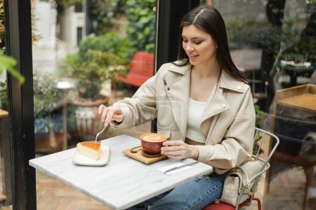 glückliche Frau mit langen Haaren, die in Lederjacke neben Fenster und Bistrotisch sitzt und eine Tasse Cappuccino und Gabel über Käsekuchen in einem modernen Café in Istanbul hält 