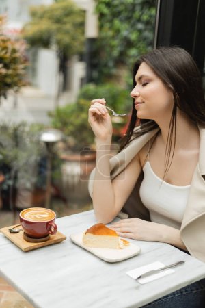 jeune femme aux cheveux longs tenant fourchette près du gâteau au fromage à côté de la tasse de cappuccino sur la table de bistrot tout en étant assis dans une veste en cuir près de la fenêtre à l'intérieur du café moderne à Istanbul 