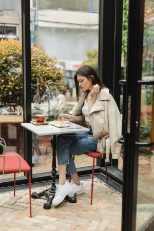 full length of young woman with long hair holding fork near cheesecake and cup of cappuccino on bistro table while sitting in leather jacket next window inside of modern cafe in Istanbul 