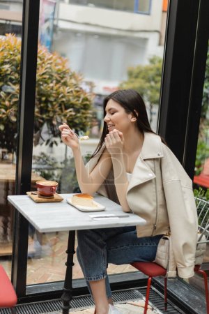 glückliche junge Frau mit langen Haaren, die Käsekuchen in der Nähe einer Tasse Cappuccino mit Kaffeekunst auf einem Bistrotisch isst, während sie in Lederjacke neben dem Fenster eines modernen Cafés in Istanbul sitzt 