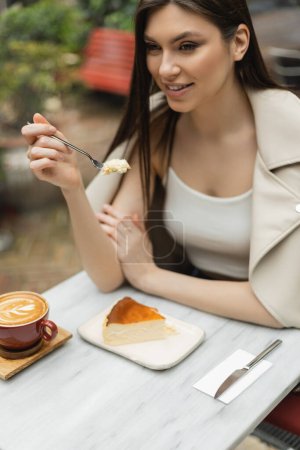 Foto de Mujer sonriente con el pelo largo sosteniendo tenedor con pastel de queso al lado de la taza de capuchino con el arte del café en la mesa del bistro mientras está sentado dentro de la cafetería moderna en Estambul - Imagen libre de derechos