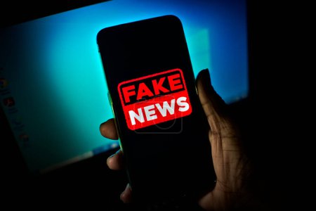 Hand holds a smartphone where the phrase fake news appears on the screen. Image with news concept and low light