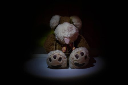 Photo for Stuffed animal illuminated by spotlight in a dark room. Child abuse and violence concept. Sad dramatic mood for negative themes such as bullying at school, child abuse, pedophilia, traumatic childhood or kidnap. - Royalty Free Image