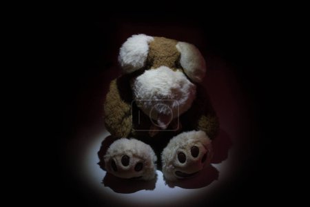 Photo for Stuffed animal illuminated by spotlight in a dark room. Child abuse and violence concept. Sad dramatic mood for negative themes such as bullying at school, child abuse, pedophilia, traumatic childhood or kidnap. - Royalty Free Image