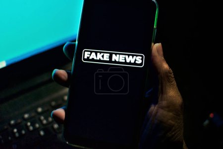 Hand tient un smartphone où l'expression fake news apparaît à l'écran. Image avec concept de nouvelles et faible luminosité