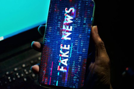 Hand holds a smartphone where the phrase fake news appears on the screen. Image with news concept and low light