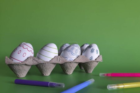 Photo for Tray with Easter eggs and markers. Copy space. Preparing for holiday with children. Set for Easter decor - blanks of eggs with pattern and colored felt tip pens on green background. Place for text. - Royalty Free Image