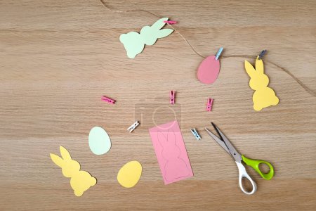Photo for Creating decorative garland for Easter. Flat lay. Crafts for children and adults. Multi colored paper rabbits and eggs on rope, clippers, scissors on wooden background. Top view. Happy Easter concept. - Royalty Free Image