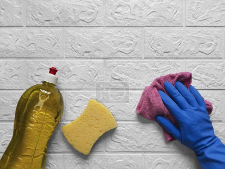 Photo for Hand with microfiber cloth. Routine work in kitchen.  Hand in blue rubber glove holds and wipes kitchen white brick wall with pink cleaning washcloth. Flat lay. Cleaning process. Copy space. - Royalty Free Image