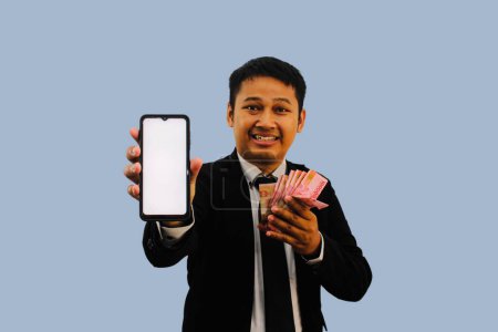 Photo for Adult Asian man smiling happy while showing blank mobile phone screen and holding paper money - Royalty Free Image