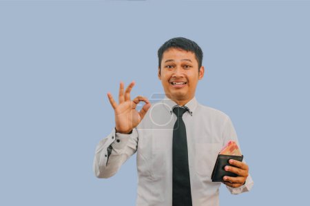 Erwachsener asiatischer Mann lächelt, während er Geldbörse mit Geld in der Hand hält und OK Fingerzeichen gibt