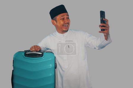 musulmán asiático hombre llevando maleta mostrando feliz expresión al mirar su teléfono