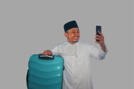 Moslem Asian man carrying suitcase showing happy expression when looking his phone