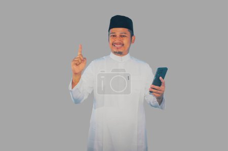 Téléchargez les photos : Musulman asiatique homme souriant heureux tout en tenant téléphone mobile et pointant vers le côté supérieur - en image libre de droit