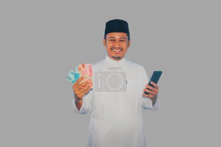 Photo for Moslem Asian man smiling happy while holding mobile phone and money - Royalty Free Image