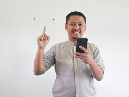 Adult Asian man smiling and pointing one hand up while holding mobile phone