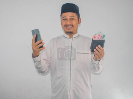 Téléchargez les photos : Asiatique musulman sourire heureux tout en tenant téléphone portable et montrant de l'argent de papier de son portefeuille - en image libre de droit