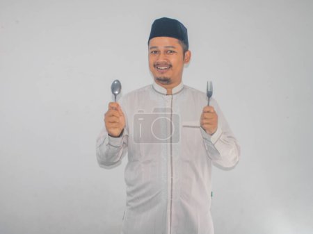 Moslem Asian man smiling happy with hand holding dinning spoon and fork