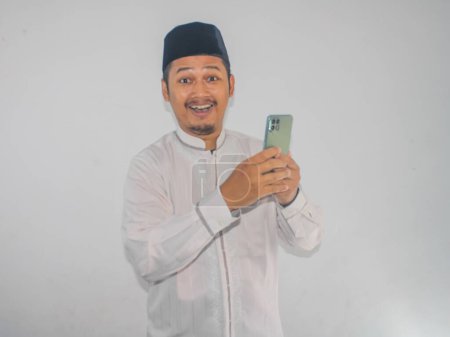 Photo for Moslem Asian man showing wow expression while holding his mobile phone - Royalty Free Image