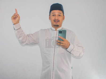 Photo for Moslem Asian man shocked while holding mobile phone and pointing to the up side - Royalty Free Image
