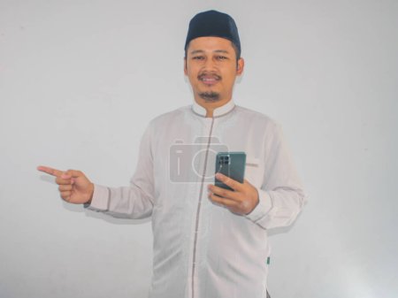 Photo for Moslem Asian man smiling happy while holding mobile phone and pointing to the right side - Royalty Free Image